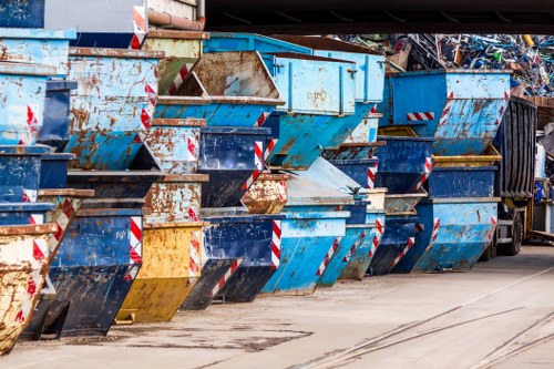 A row of large industrial metal skips and containers, primarily painted in blue with some showing signs of rust and wear. The skips are positioned side by side on a paved surface, with some partially filled with mixed waste materials. The containers are arranged outdoors, possibly on a commercial waste disposal site, with a few leaning slightly forward and others stacked at slight angles. In the background, there are additional waste bins and a portion of a trailer or truck visible. The ground beneath the skips is concrete with visible tire marks and stains, indicating frequent vehicle movement. The overall scene reflects a typical waste collection area where commercial rubbish is temporarily stored before removal. The presence of rust, dirt, and weathered finishes suggests the area services regular industrial or business waste removal, aligning with the services of Commercial Waste Removal Regents Park in the London boroughs, providing efficient rubbish disposal solutions in the local area near NW1 or NW8 postcode regions.
