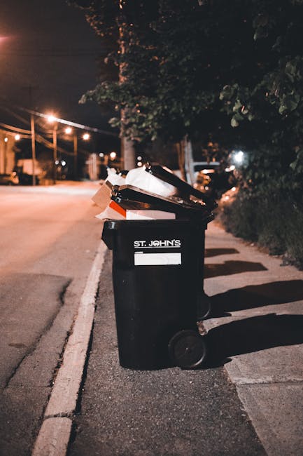 A black wheelie bin marked with the label 'St. John's' placed on the pavement alongside a curb in an urban street at night. The bin is filled with various waste items, including cardboard and plastic 